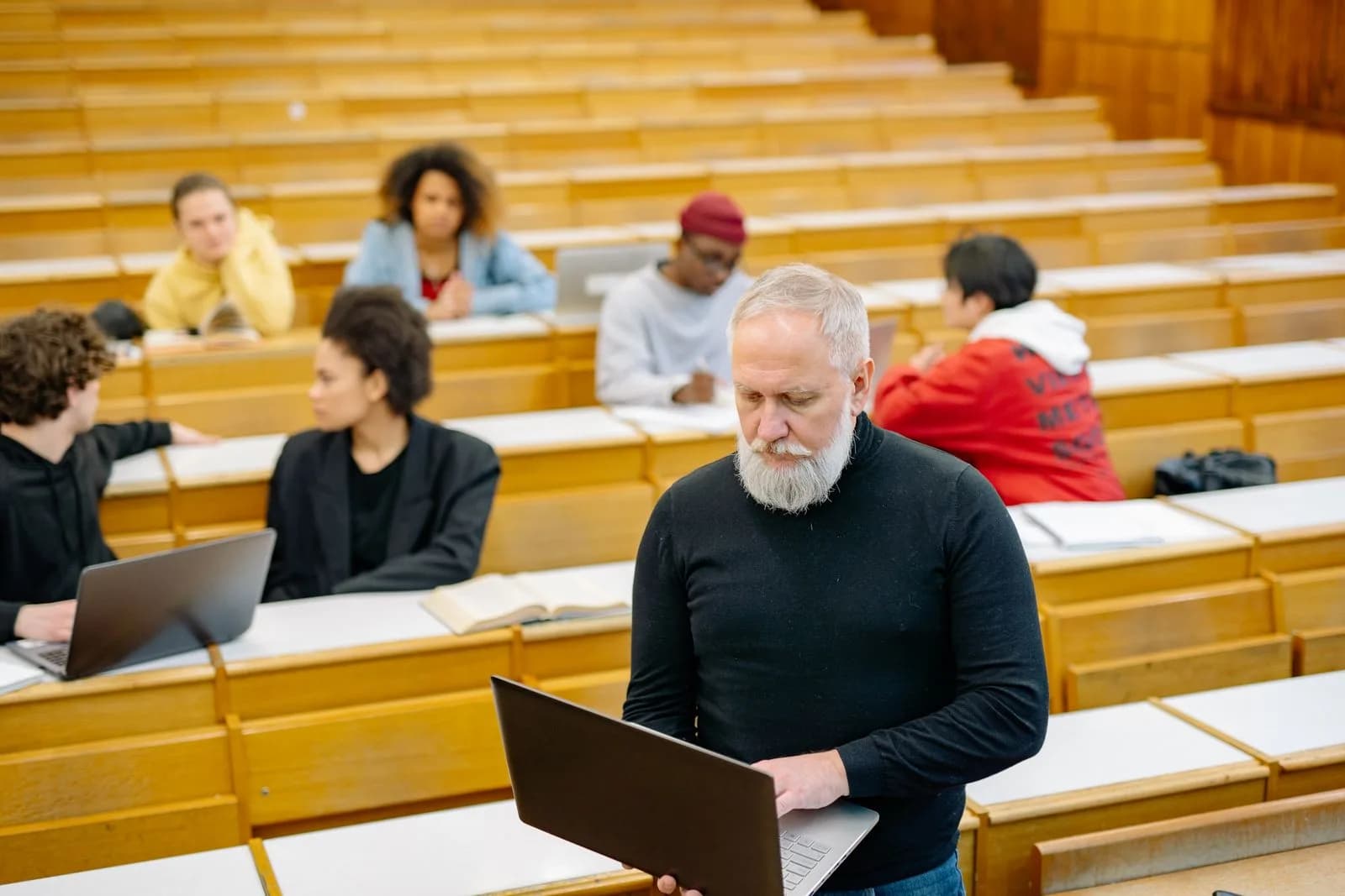 An educator working on his laptop in a lecture hall, students at their desks behind him